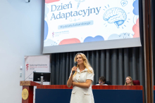 Kobieta stoi na scenie i przemawia do uczestników wydarzenia. W tle widoczny jest ekran z grafiką Dnia Adaptacyjnego.