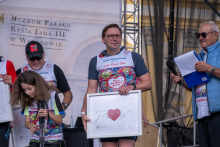A participant stands on stage holding a framed artwork featuring a heart motif. Other participants and a presenter with a microphone are nearby.