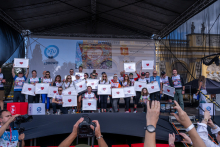 A group of participants stands on stage presenting framed artworks featuring a heart motif. Photographers and media representatives are gathered in front of the stage.