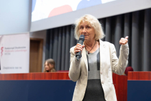 Kobieta stoi na scenie i przemawia do uczestników wydarzenia, trzymając mikrofon w dłoni. W tle znajduje się stół prezydialny oraz ekran prezentacyjny.