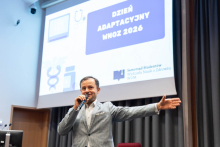 Mężczyzna stojący na scenie prowadzi prezentację i gestykuluje wolną ręką. W tle znajduje się ekran z grafiką Dnia Adaptacyjnego WNoZ 2026.