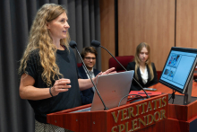 Kobieta, Magdalena Łazarewicz, prowadzi prezentację przy mównicy. Na monitorze widoczny jest slajd prezentacji, a przy stole prezydialnym siedzą dwie osoby.
