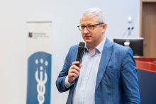 Mężczyzna w niebieskiej marynarce przemawia do uczestników, trzymając mikrofon w dłoni.