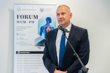 Osoba przemawia przy mikrofonie podczas Forum WUM-PW. W tle znajduje się roll-up informacyjny wydarzenia.
