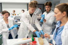 Grupa uczestników wykonuje ćwiczenia mikrobiologiczne przy stole laboratoryjnym pod opieką prowadzącej.