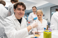 Osoby w fartuchach laboratoryjnych pracują przy stanowisku laboratoryjnym. Na pierwszym planie widoczna jest płytka Petriego oraz narzędzia do badań mikrobiologicznych.