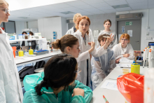 Prowadząca zajęcia prezentuje próbkę laboratoryjną grupie uczestników zgromadzonych wokół stołu roboczego w laboratorium.