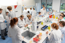 Widok ogólny laboratorium, w którym grupa uczestników wykonuje ćwiczenia przy kilku stanowiskach wyposażonych w sprzęt laboratoryjny.