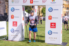A participant using Nordic walking poles passes beneath an event arch during the “Run for New Life” march. Other participants are visible nearby.