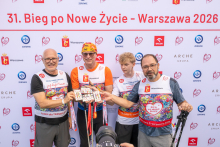Four participants pose in front of the “31st Run for New Life – Warsaw 2026” backdrop and present commemorative medals. One participant points toward the medals held by the group.