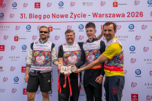 Four people pose for a photo in front of the backdrop of the “31st Run for New Life – Warsaw 2026” event. The participants display commemorative medals.