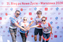 Four people stand in front of the event backdrop and place their hands together. Commemorative medals hang around their necks.