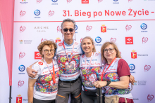 Four people stand together in front of the backdrop of the “31st Run for New Life – Warsaw 2026” event. They wear medals earned during the event.