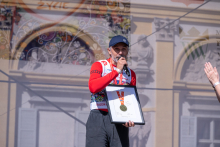 One person speaks on stage while holding a microphone and a framed commemorative award with a medal. A palace-themed backdrop is visible in the background.
