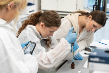Trzy osoby pracują przy stole laboratoryjnym. Jedna osoba pobiera próbkę za pomocą pipety automatycznej.