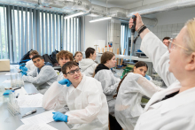 Prowadząca prezentuje sposób użycia pipety automatycznej. Kilkanaście osób obserwuje pokaz przy stanowiskach laboratoryjnych.