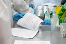 Osoba w rękawiczkach przegląda instrukcję ćwiczenia laboratoryjnego. Obok stoją probówki i stojaki laboratoryjne.