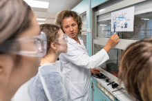 Prowadząca wyjaśnia działanie aparatury laboratoryjnej, wskazując element schematu zawieszonego obok stanowiska.