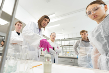 Grupa uczestników obserwuje pokaz doświadczenia laboratoryjnego przy stanowisku z naczyniami i odczynnikami.