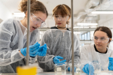 Trzy osoby w okularach ochronnych analizują próbkę podczas ćwiczenia laboratoryjnego przy stanowisku z naczyniami laboratoryjnymi.