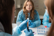 Osoba w rękawiczkach trzyma probówkę z próbką krwi. Kilka osób uczestniczy w zajęciach laboratoryjnych przy stole.