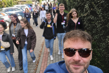 Grupa uczestników spaceru edukacyjnego porusza się chodnikiem wzdłuż budynku. Jedna osoba wykonuje zdjęcie obejmujące całą grupę.