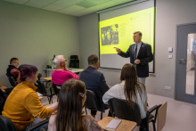A man is giving a lecture to participants seated in a training room. He is pointing toward a presentation displayed on a screen.