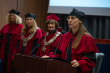 A person wearing a red academic gown and a black mortarboard speaks at a lectern with two microphones, holding an open folder. In the background stand three people in red academic gowns and mortarboards, two of them wearing ceremonial chains.
