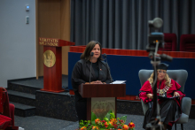 A woman speaks at the lectern during the event. A person wearing an academic gown can be seen seated nearby.