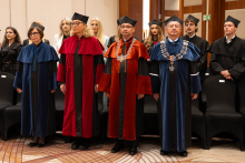 Representatives of the university authorities, wearing ceremonial academic robes and insignia, stand in the front row, with other ceremony participants behind them.