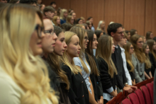A large number of ceremony attendees stand among rows of red chairs in the auditorium. In the foreground, a group of people stand side by side, facing in the same direction.