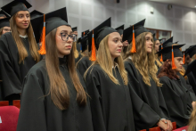 A group of graduates in black academic gowns and mortarboards with orange tassels stand in rows in the auditorium during the ceremony.