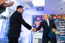 Two people shake hands in a room equipped with medical equipment. In the background, there is a Great Orchestra of Christmas Charity graphic and an information screen.