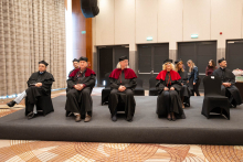 People in black and red academic attire sit in a row on a raised platform during the ceremony.