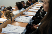 Several dozen people are seated in a lecture hall. Folded white coats are placed on the desks in front of them.