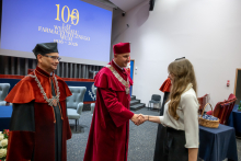 Osoba w czerwonej todze akademickiej podaje rękę uczestnikowi uroczystości. W tle widoczny ekran z napisem „100 lat Wydziału Farmaceutycznego WUM 1926–2026”.