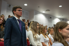 Ceremony participants stand in rows in an auditorium. In the foreground, a person wearing a dark suit and tie stands alongside several people dressed in light and dark clothing.
