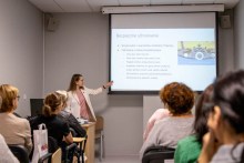 A woman is giving a presentation in a training room. A virtual reality headset is placed on the desk beside her.