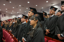 A group of graduates in black academic gowns and mortarboards with orange tassels stand in rows in the auditorium during the ceremony.