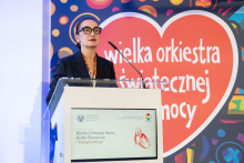 The hospital director speaks at a lectern during the ceremony. In the background, the Great Orchestra of Christmas Charity logo and a screen displaying information about the clinic can be seen.