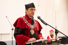 A person in red academic robes and a ceremonial chain speaks into a microphone at the lectern during an academic ceremony.