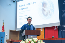 A person wearing a blue academic gown and black cap speaks at a lectern. A presentation slide titled “Before a human is born. Contemporary possibilities of fetal therapy” with an illustration of a fetus is displayed on the screen in the background.