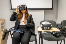 A woman is seated on a chair using a virtual reality headset. Another headset is placed on a nearby chair.