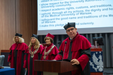 A person wearing a red academic gown and a black mortarboard speaks at a lectern with two microphones, holding an open folder. In the background stand three people in red academic gowns and mortarboards, two of them wearing ceremonial chains.
