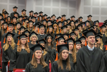 A group of graduates in black academic gowns and mortarboards with orange tassels stand in rows in the auditorium during the ceremony.
