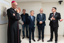A group of six people stand in a hospital room during a presentation. One person is speaking while the others listen.