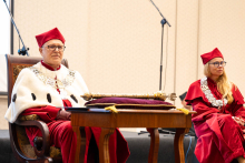 Two people in ceremonial red-and-white academic robes and insignia sit on stage; an ornate academic mace rests on the table in front of them.
