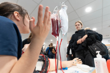 Two people talk at a station equipped with a medical procedure simulation kit. Bags of fluids are suspended from a stand and connected by tubing to a model arm.