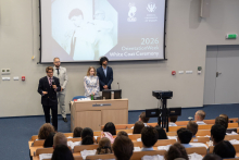 Four people are standing at the front of the auditorium during the White Coat Ceremony. One person is addressing the participants gathered in the auditorium.