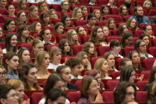A large group of ceremony participants sit in rows of red seats in an auditorium. Some of the participants are applauding.